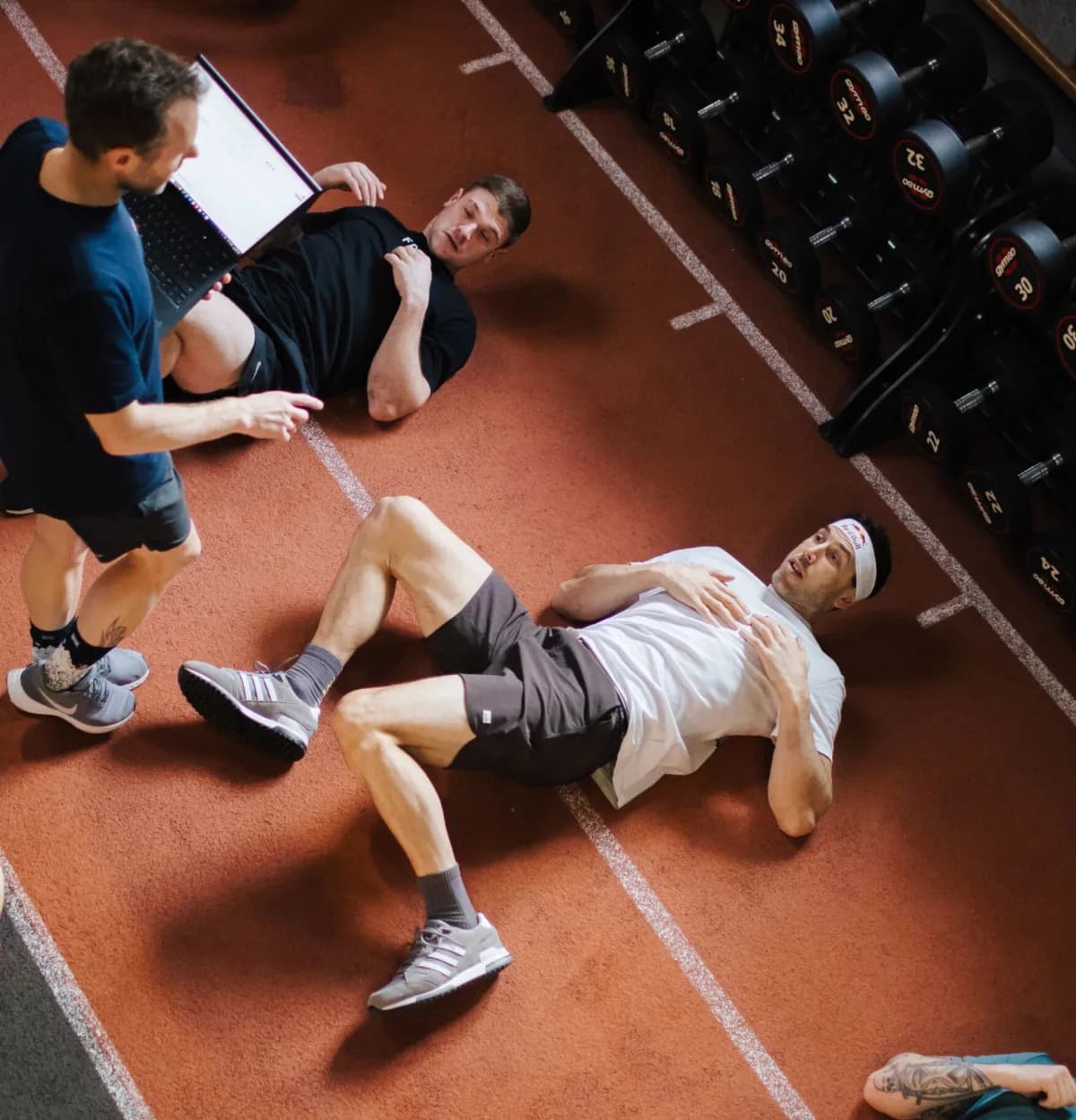 Coach reviewing data on a laptop while athletes train on an indoor track