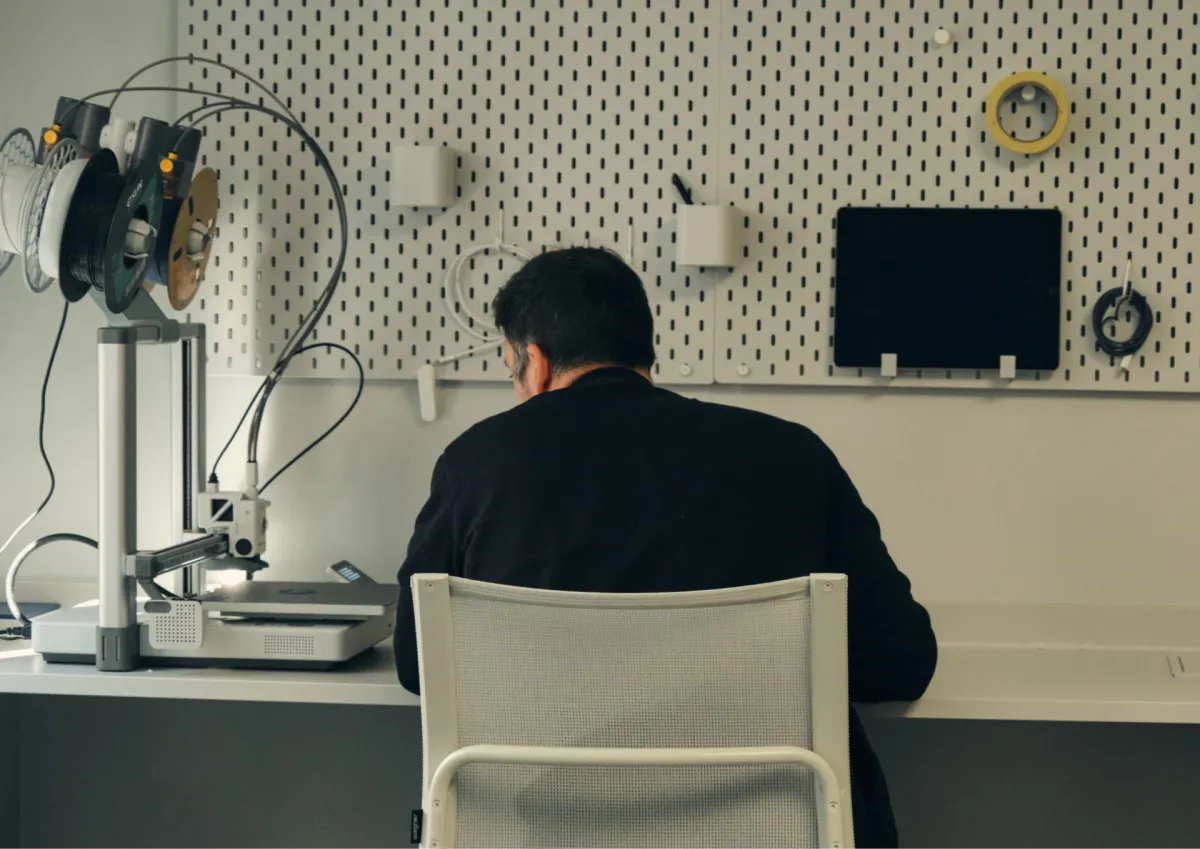 Person sitting at a workbench with a 3D printer and tools organized on a pegboard wall