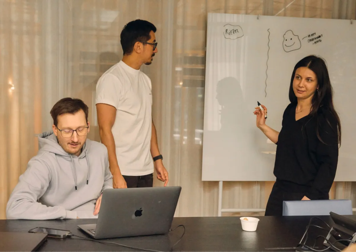 Three people collaborating in front of a whiteboard, with one person seated at a table with a laptop
