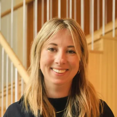 Person with shoulder-length blonde hair, wearing a black top, standing in front of a wooden staircase