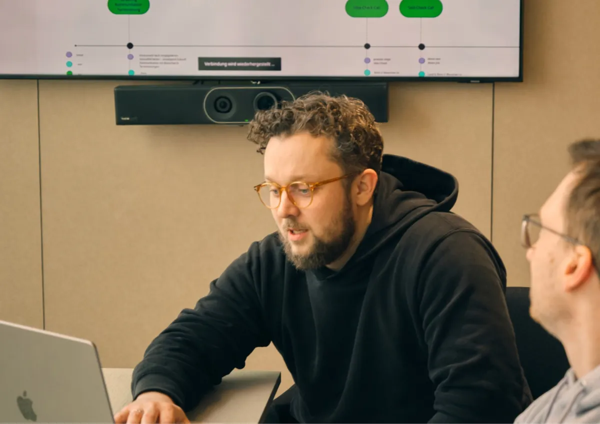 Two people having a discussion in a meeting room with a laptop and a large screen displaying a diagram