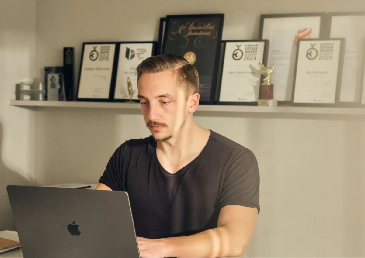Person working at a desk with a laptop, with several awards and certificates displayed on a shelf in the background